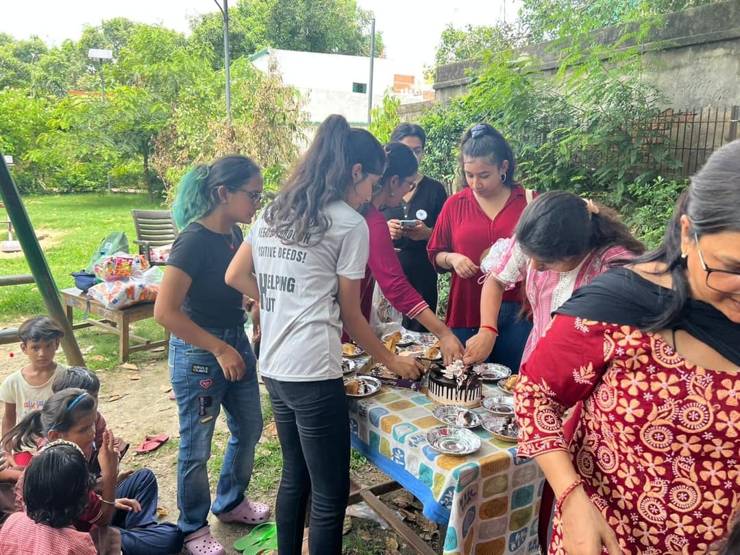 Helping Hut volunteers in NGO t-shirts plate slices of chocolate cake at an outdoor table while children wait nearby during a park celebration.