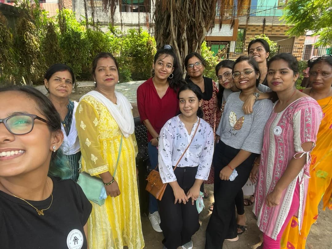 Helping Hut volunteers and community women smile together in a group selfie under a leafy tree after a celebration.