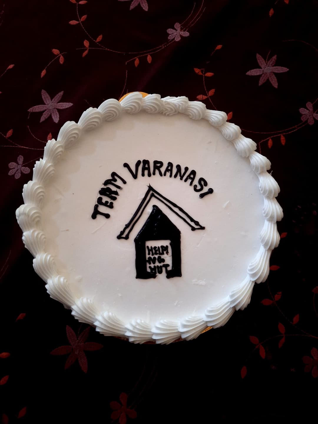 A white frosted cake decorated with the Helping Hut house logo and the words Team Varanasi piped in chocolate icing.