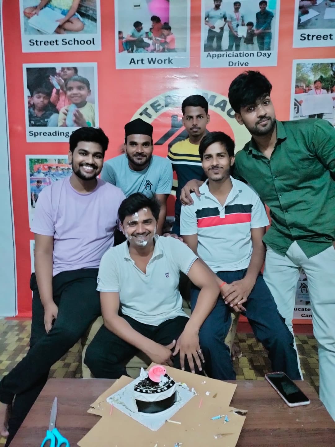 Six Helping Hut volunteers pose around a birthday cake indoors, one with frosting on his face, in front of a board showcasing the NGO's activities.