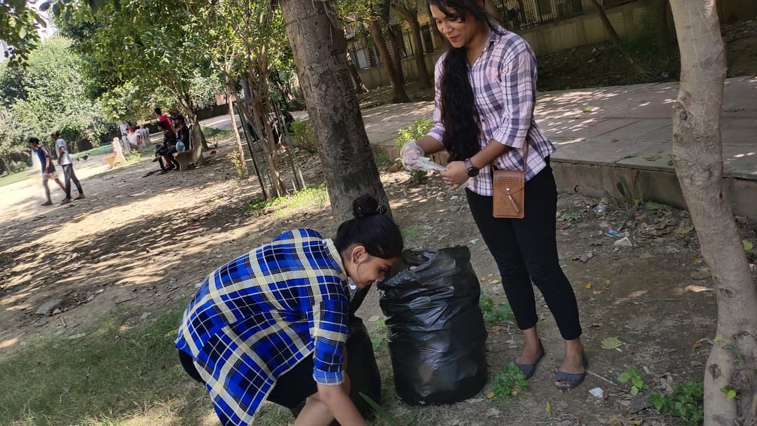 A Helping Hut volunteer bends to pick litter from the grass while another volunteer holds open a black garbage bag under park trees.