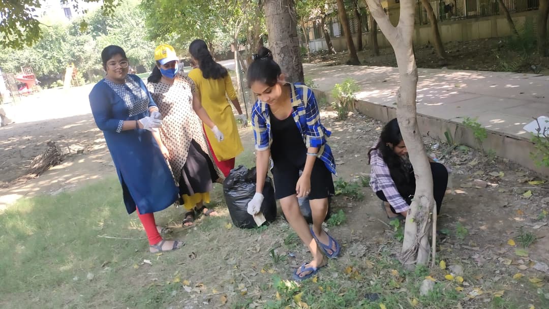 A group of gloved Helping Hut volunteers collects litter from a grassy park, gathering the waste into a shared black garbage bag.