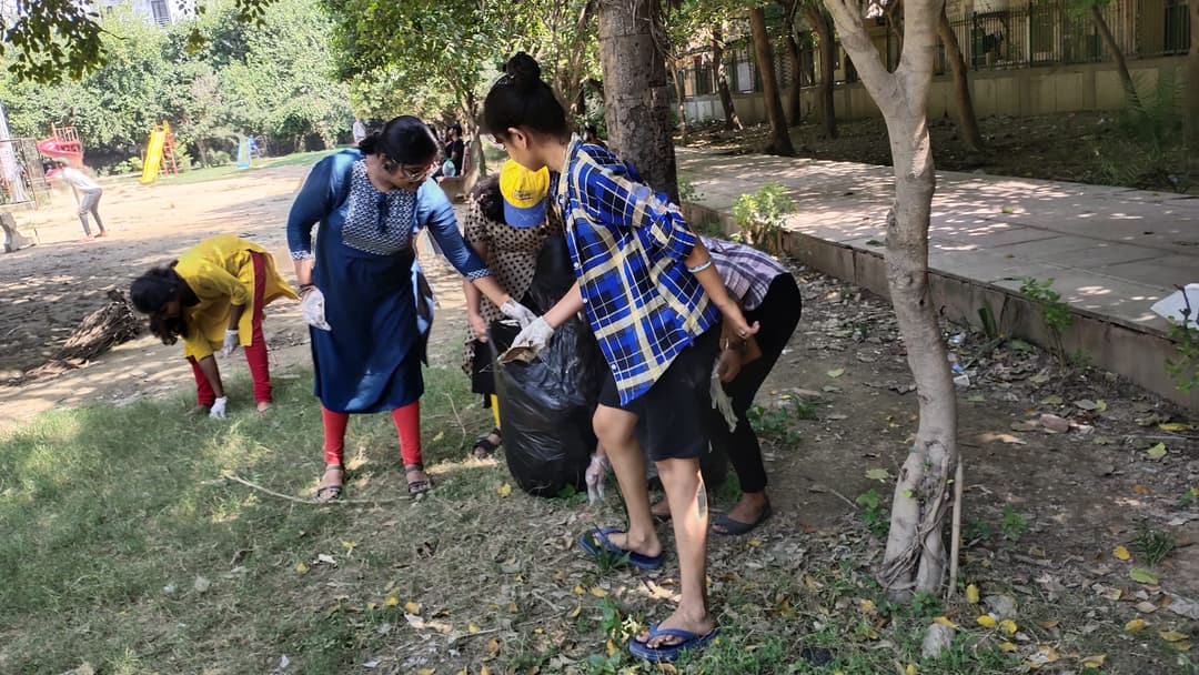 Helping Hut volunteers gather around a black garbage bag while picking up litter beneath trees in a park.