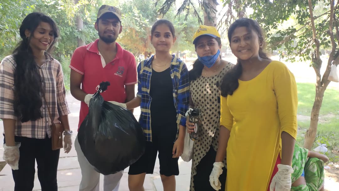 Five smiling Helping Hut volunteers wearing gloves pose with a filled garbage bag after completing a park cleanliness drive.