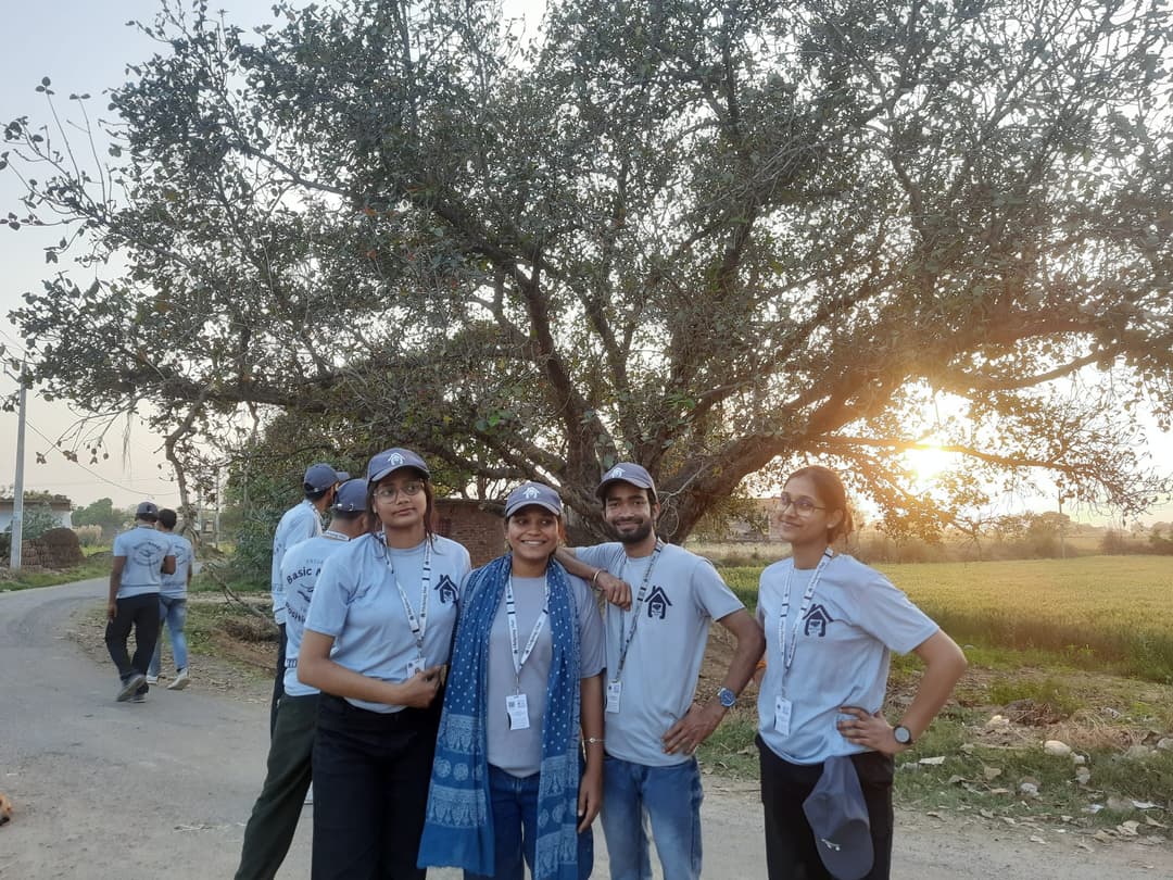Four Helping Hut volunteers in team t-shirts, caps and ID lanyards pose on a village road at sunset, with a large tree and green fields behind them.