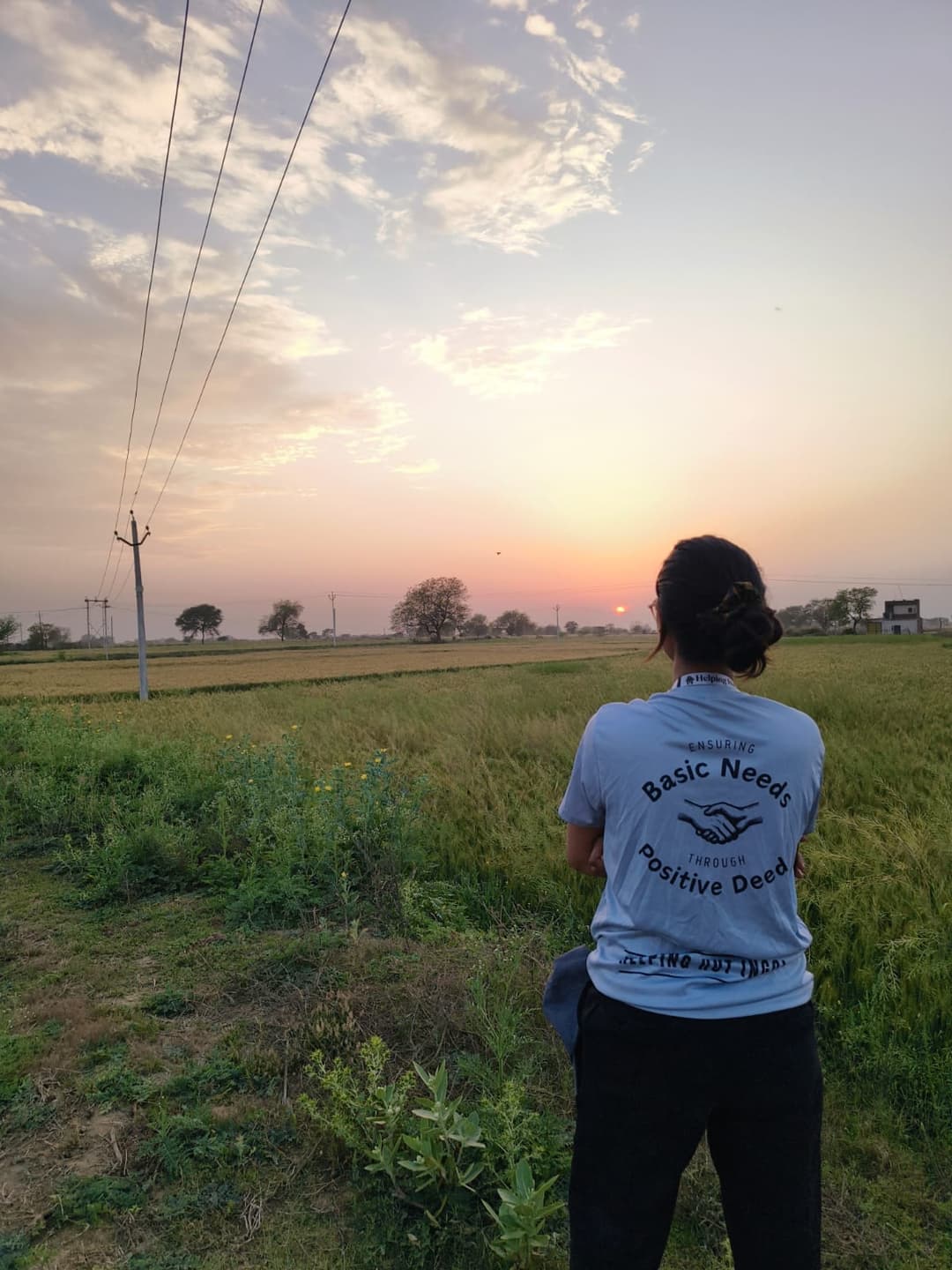 A Helping Hut volunteer in the NGO t-shirt reading Ensuring Basic Needs Through Positive Deeds watches the sun set over green wheat fields at the edge of the village.