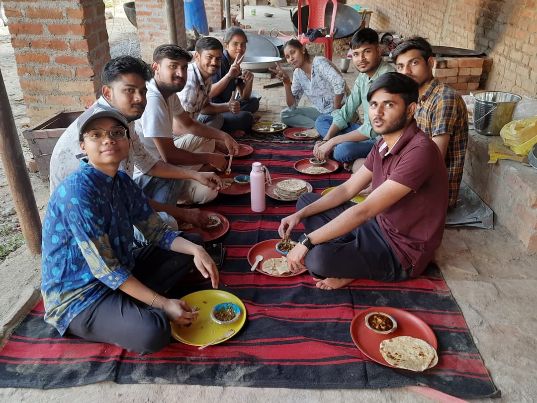 Helping Hut volunteers sit cross-legged on mats in a village courtyard kitchen, sharing a simple meal of rotis and curry from bright plastic plates.