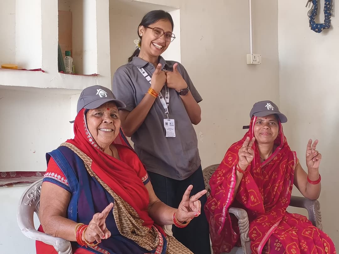 A smiling Helping Hut volunteer gives a double thumbs up between two village women in red saris who wear Helping Hut caps and flash victory signs.