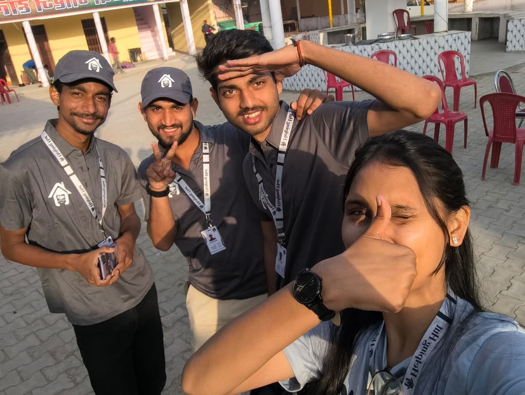 Four Helping Hut volunteers in caps and lanyards pose for a playful selfie in a paved village courtyard, one saluting and another winking with a thumbs up.