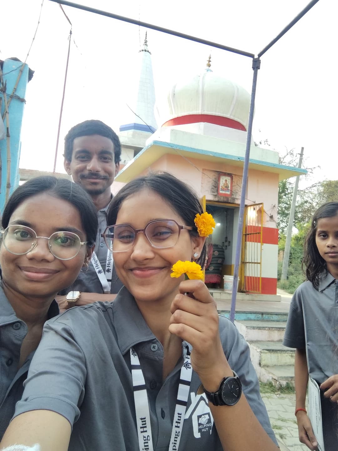 Helping Hut volunteers take a selfie holding marigold flowers outside a small village temple, joined by a local girl.