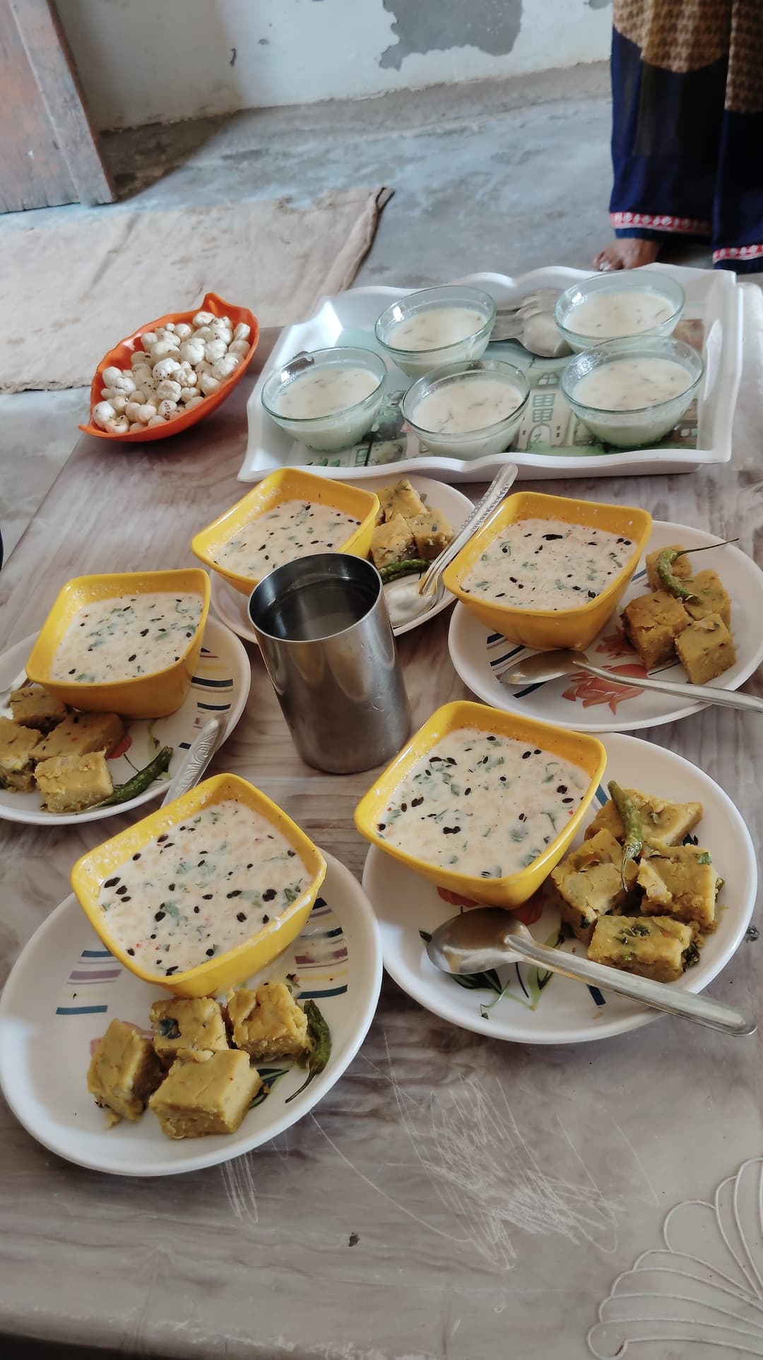 A home-cooked village breakfast laid out on a floor table: bowls of seasoned curd, plates of steamed savoury cakes with green chillies, and a dish of makhana.