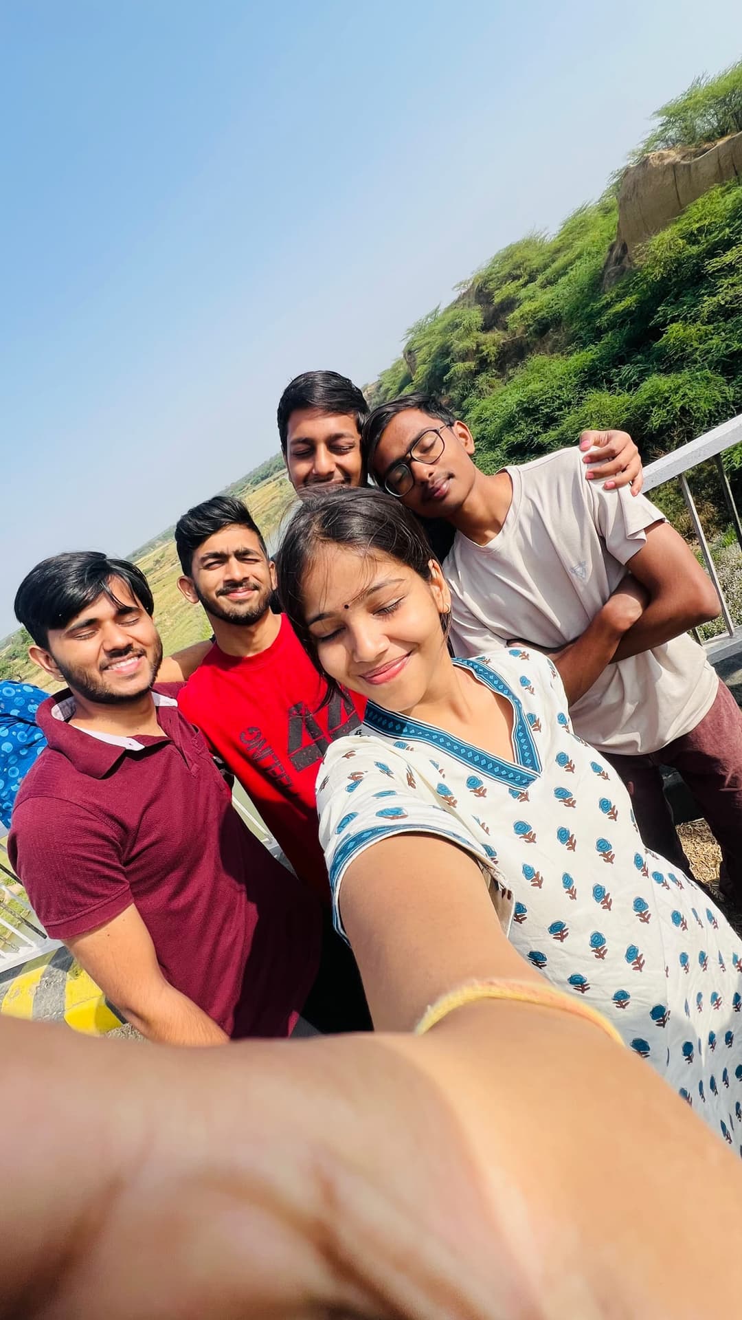 Five Helping Hut volunteers squeeze into a sunlit selfie on the river bridge, squinting and smiling against the bright morning sun.