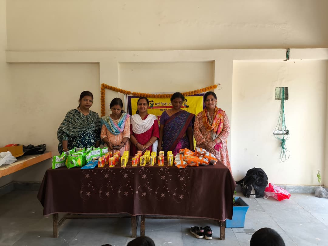 Five women stand behind a table stacked with biscuit packets, snack pouches and Frooti juice cartons arranged for distribution in a community hall decorated with a marigold-trimmed banner.