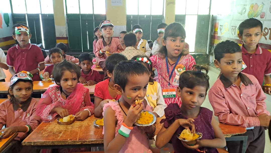 Schoolchildren wearing tricolour caps and wristbands eat distributed snacks from plates at their classroom desks during a festive celebration.