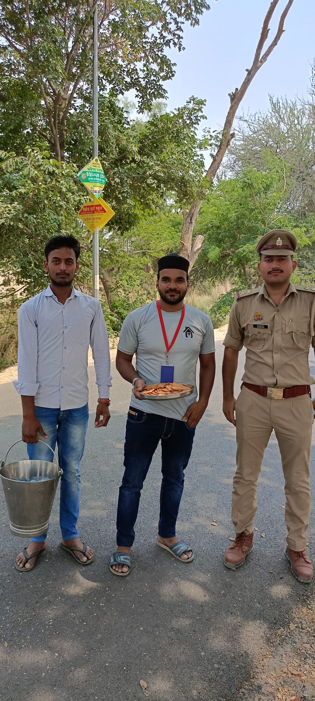 A Helping Hut volunteer with an ID lanyard holds a plate of snacks on a tree-lined roadside, standing alongside a police officer and a local resident during a summer outreach drive.