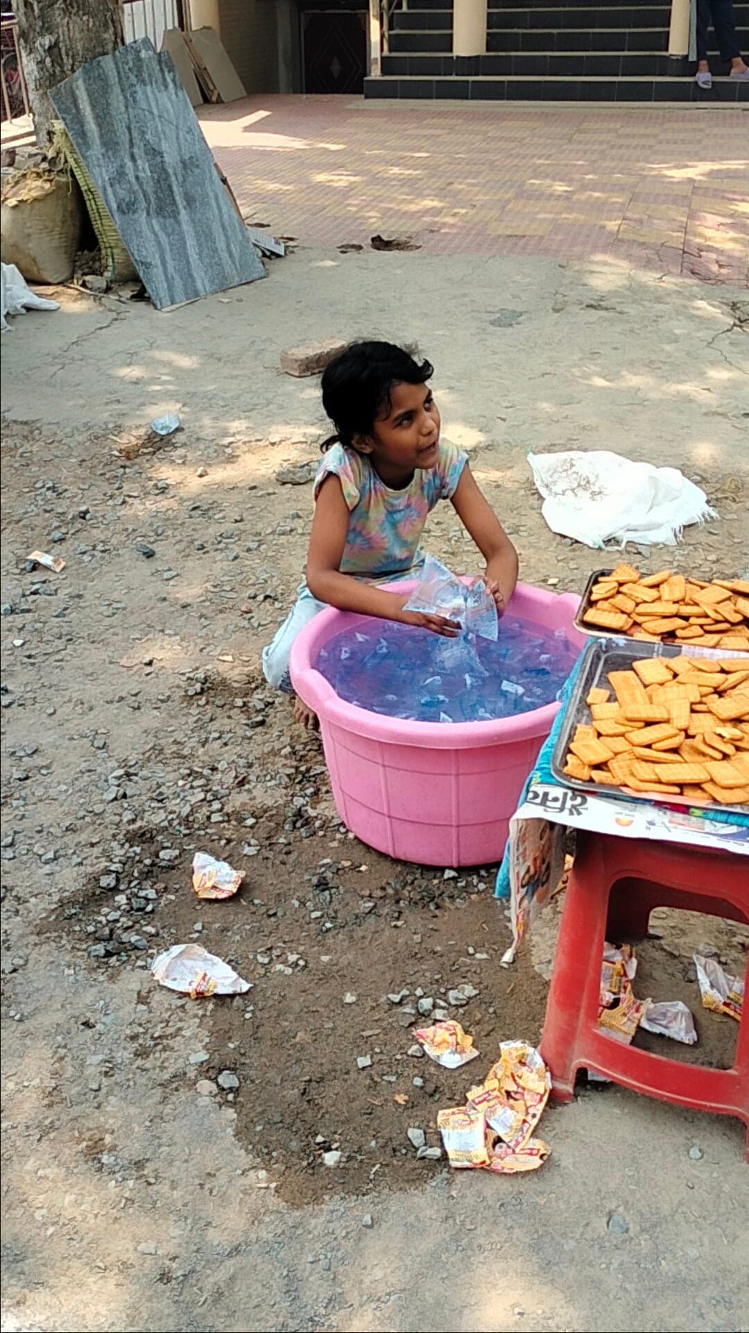 A young girl reaches into a pink tub filled with chilled water pouches next to trays of biscuits set out for a streetside distribution.