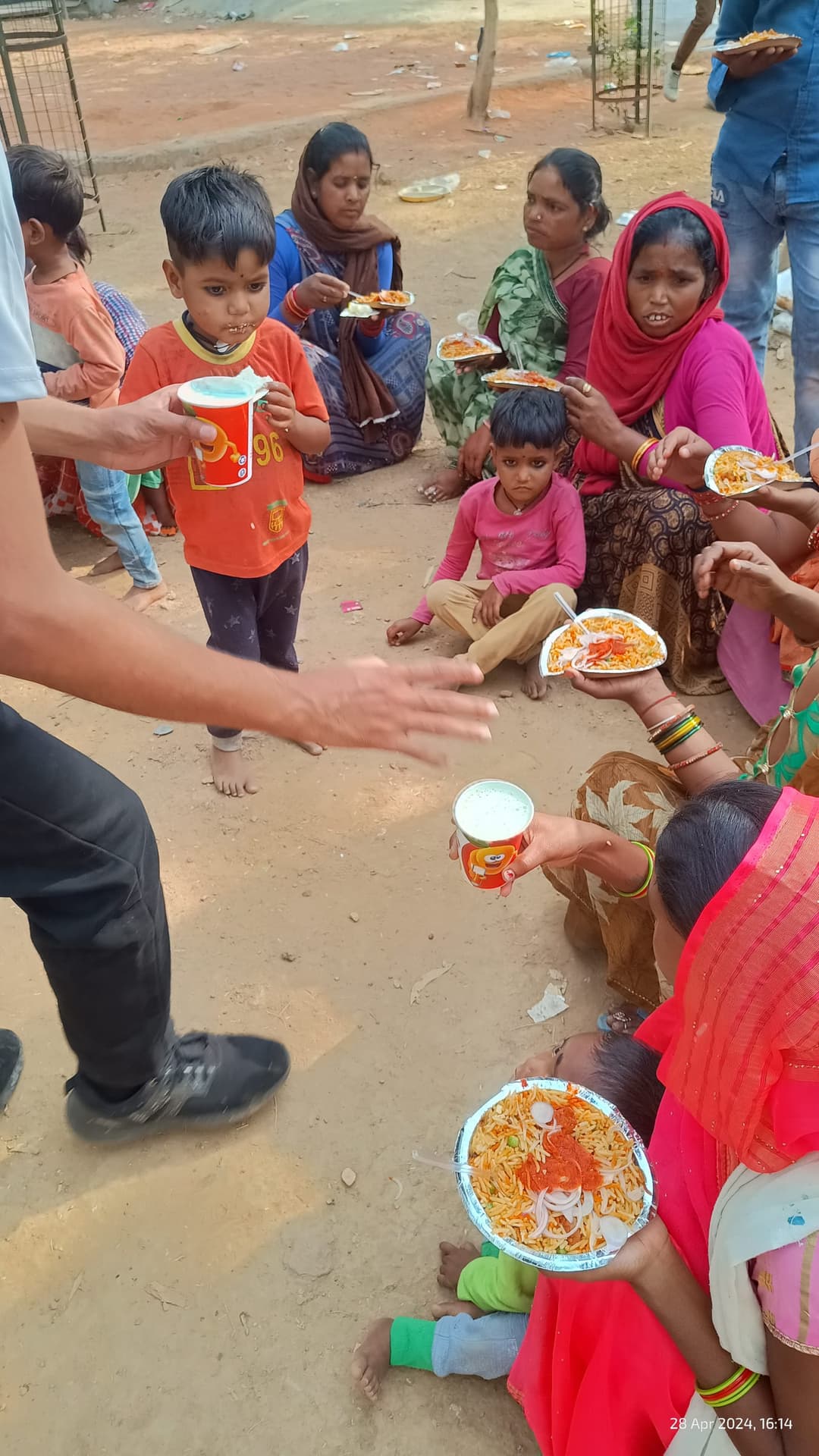 Women and children seated outdoors in a settlement hold plates of rice and cups of drink as a Helping Hut volunteer hands out servings.