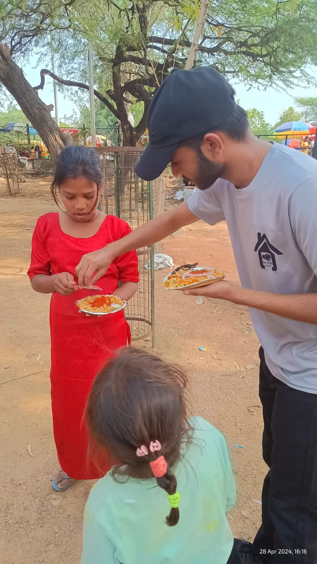 A Helping Hut volunteer bends down to hand a plate of food to a young girl in a red dress during an outdoor meal distribution.