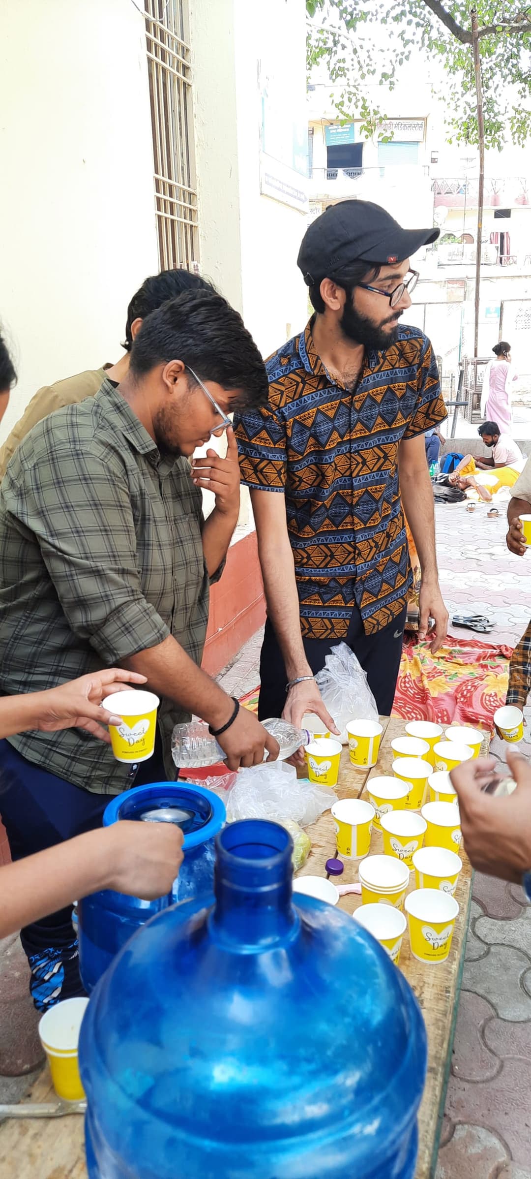 Helping Hut volunteers fill yellow cups at an outdoor table stocked with large blue water jugs while preparing a cold drink distribution.