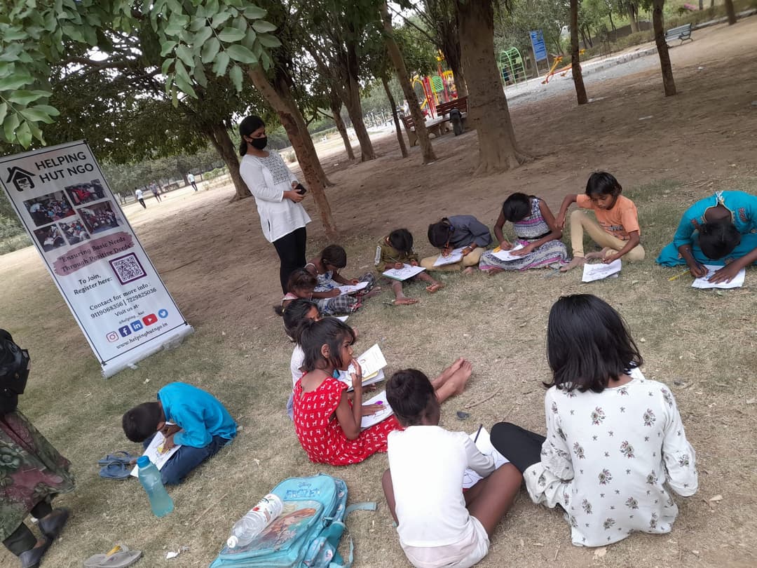 Children seated on the grass under park trees working on worksheets while a Helping Hut volunteer stands beside the NGO banner guiding the open-air class.