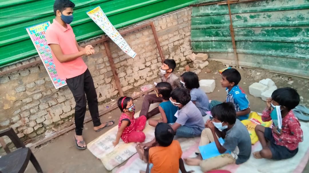 A Helping Hut volunteer teaches from colorful alphabet picture charts while children sit on mats against a brick wall during a street class.