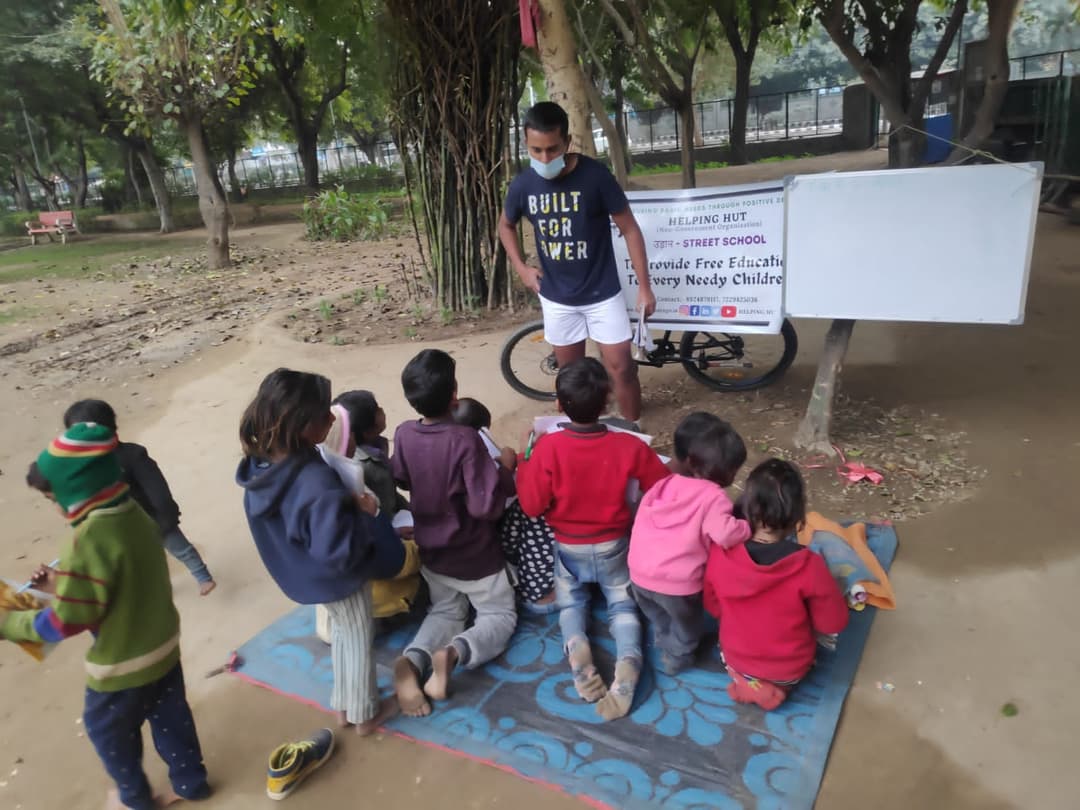 Children kneel over their notebooks on a mat in a park while a Helping Hut volunteer supervises beside the Udaan Street School board promising free education to every needy child.