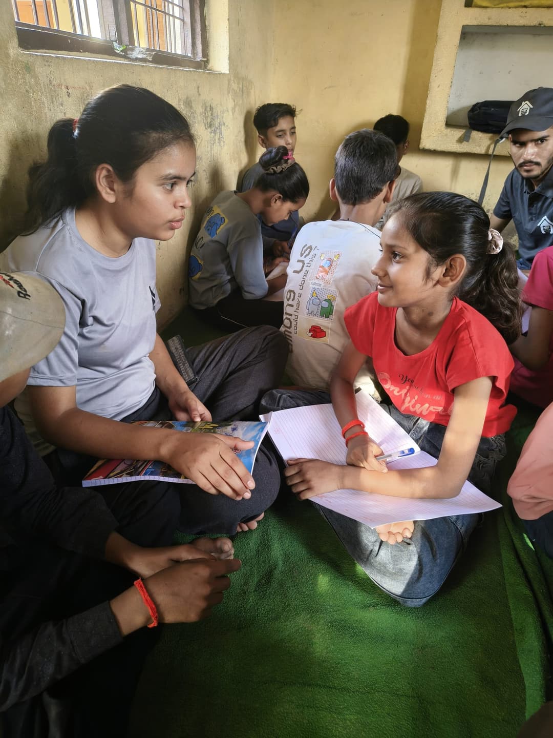 A Helping Hut volunteer sits on the classroom floor giving one-on-one guidance to a smiling girl holding her worksheet.