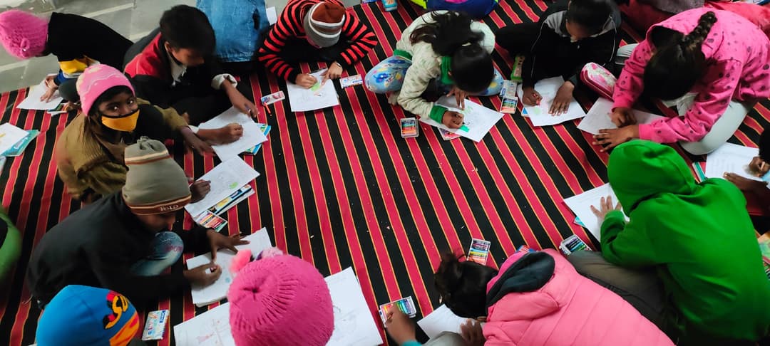 Overhead view of children in winter caps sitting in a circle on a striped mat, drawing and coloring worksheets together.