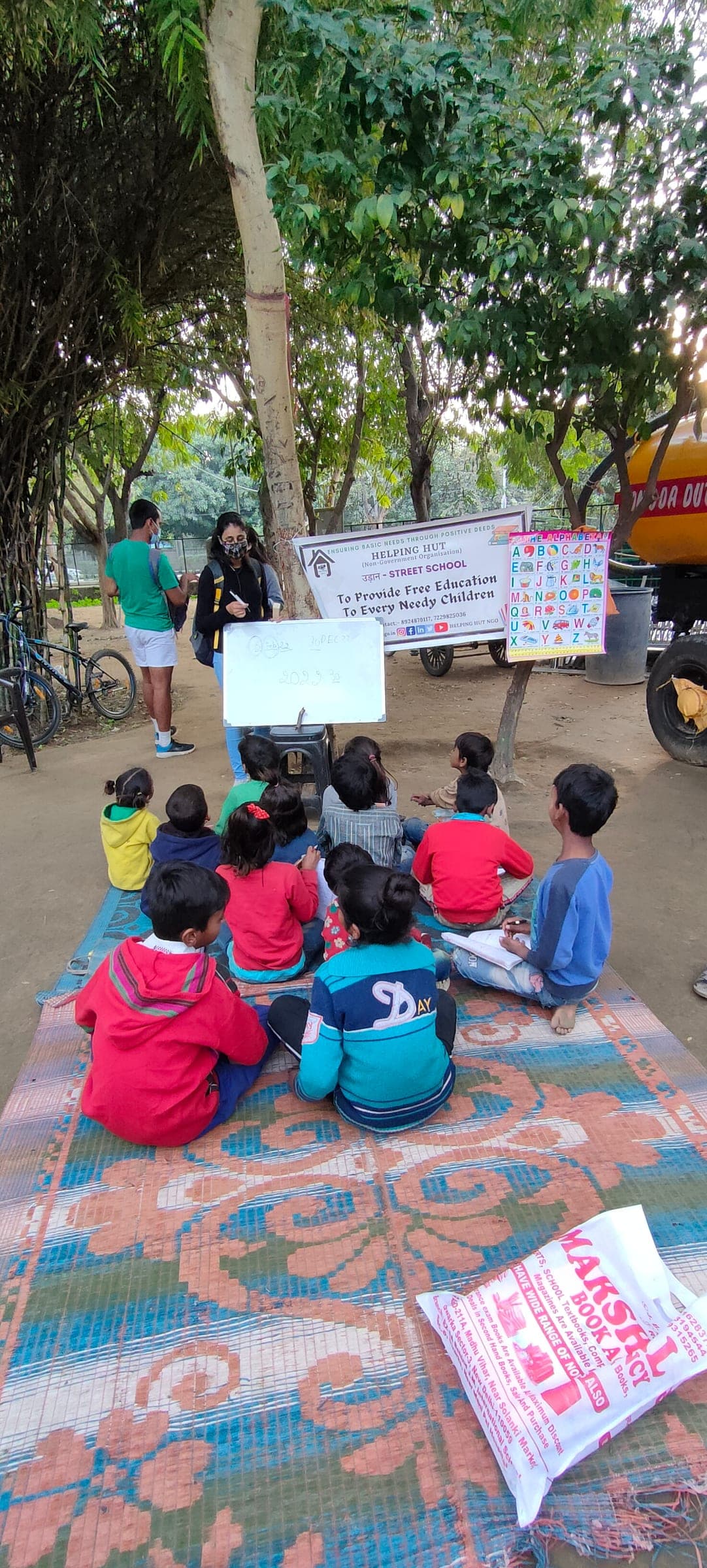 Children seated on a mat under trees follow a lesson as two Helping Hut volunteers teach with a whiteboard and alphabet chart beside the Udaan Street School banner.