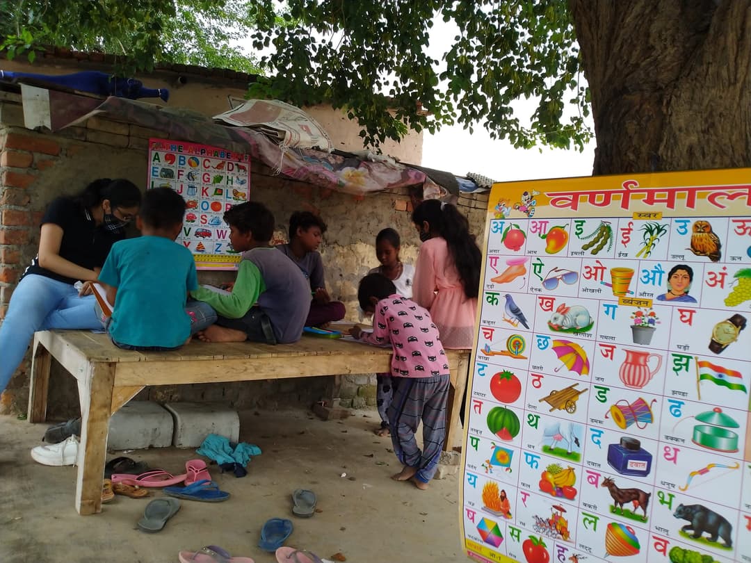 Children study at a wooden table outdoors while a Helping Hut volunteer helps them, with a large Hindi varnamala alphabet chart propped in the foreground.