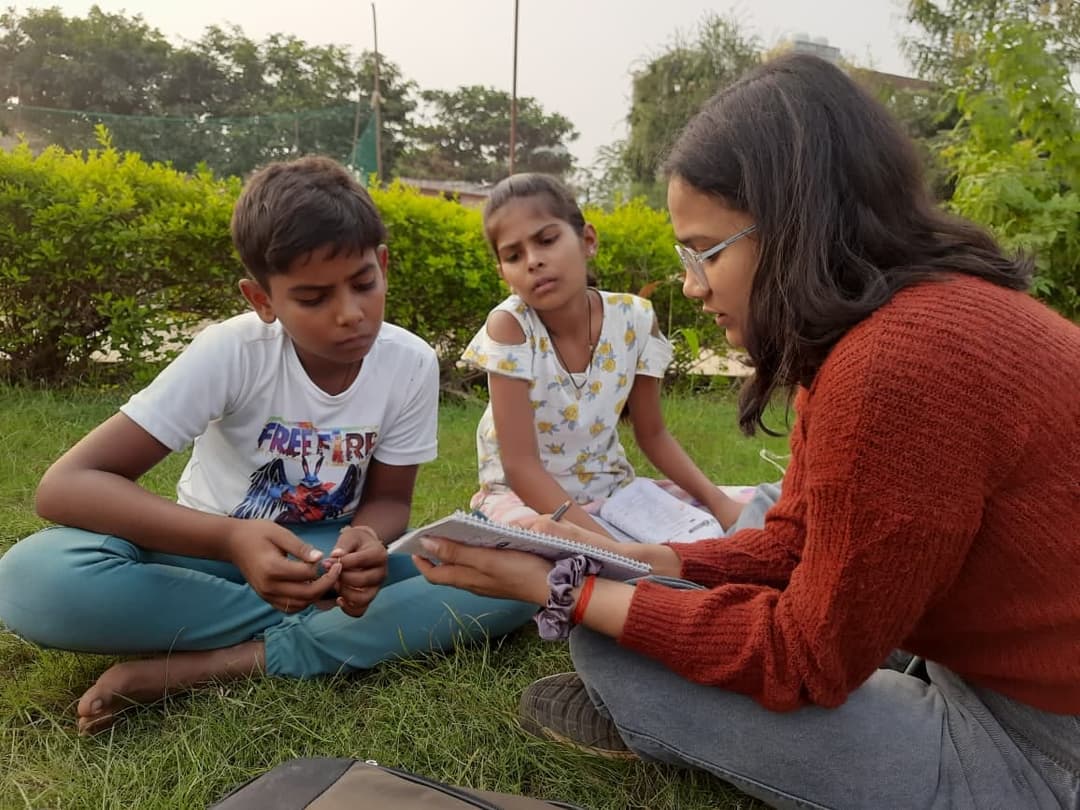 A Helping Hut volunteer sits on the grass with a notebook, working closely with a boy and a girl during an outdoor lesson.