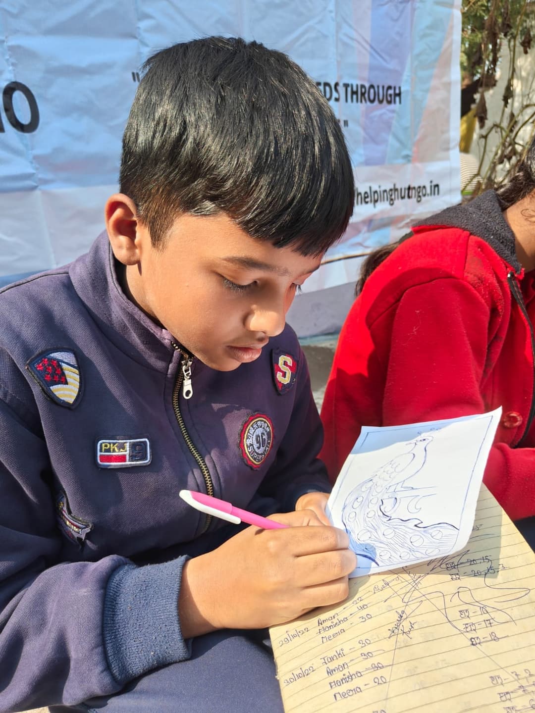 A young student concentrates on drawing a peacock with a pen at an outdoor desk, with the Helping Hut banner in the background.