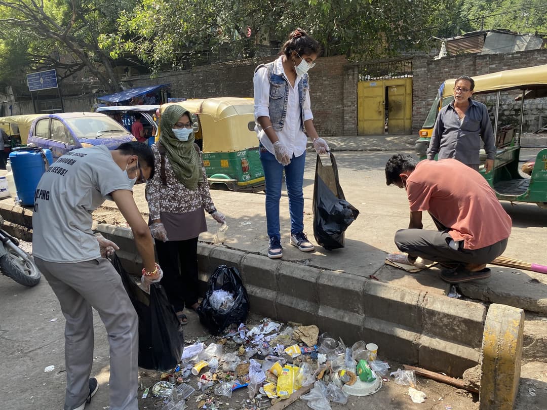 Masked Helping Hut volunteers wearing gloves pick scattered litter from a roadside curb and fill black garbage bags beside parked auto-rickshaws.