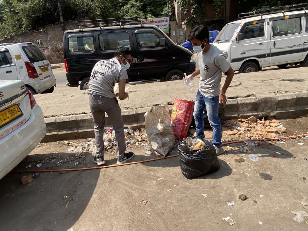 Two Helping Hut volunteers in team t-shirts and masks bag trash from a curb between parked cars during a roadside cleanliness drive.