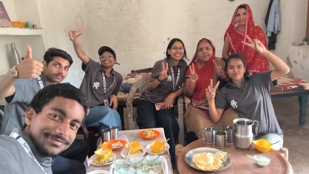 Helping Hut volunteers take a cheerful group selfie with village women in a home, with snacks and steel tumblers laid out on a table in front of them.
