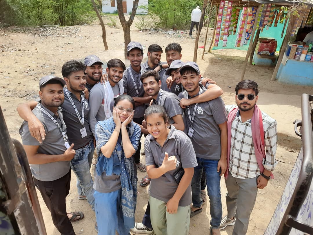 A dozen Helping Hut volunteers in grey team t-shirts and caps huddle together for a playful group photo on a village lane beside a small shop.