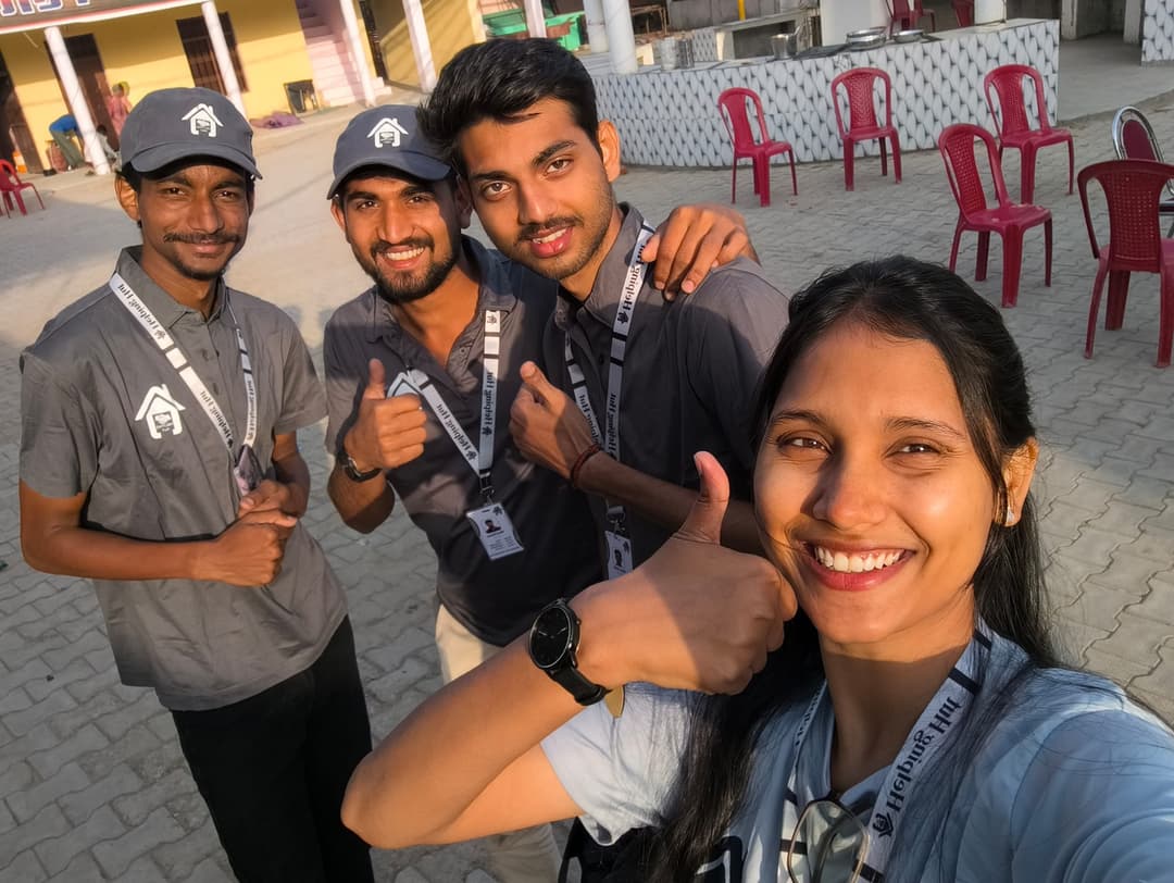 Four Helping Hut volunteers give thumbs up in a sunny selfie taken in a village courtyard with red plastic chairs in the background.