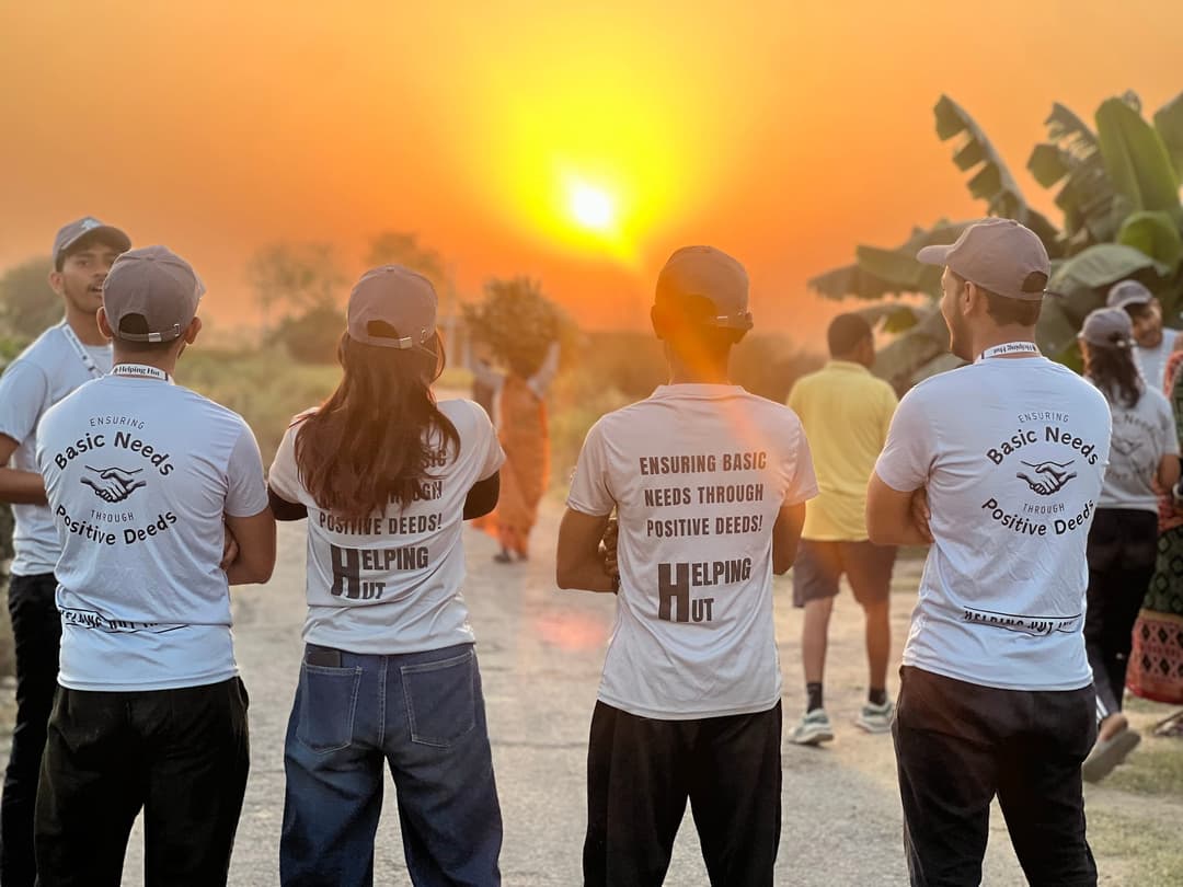 Four Helping Hut volunteers in Ensuring Basic Needs Through Positive Deeds t-shirts stand on a village road watching a glowing orange sunset, while a villager carries fodder on her head in the distance.