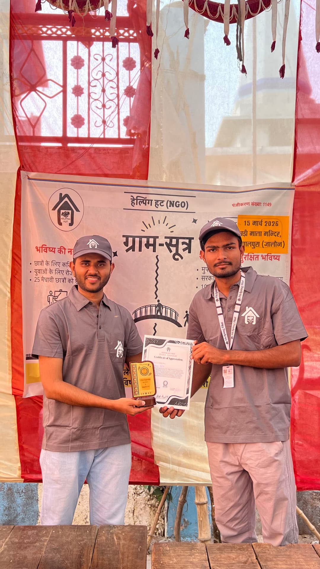Two Helping Hut volunteers hold up a memento and certificate of appreciation in front of the GramSutr campaign banner at Badi Mata Mandir, Gopalpura.