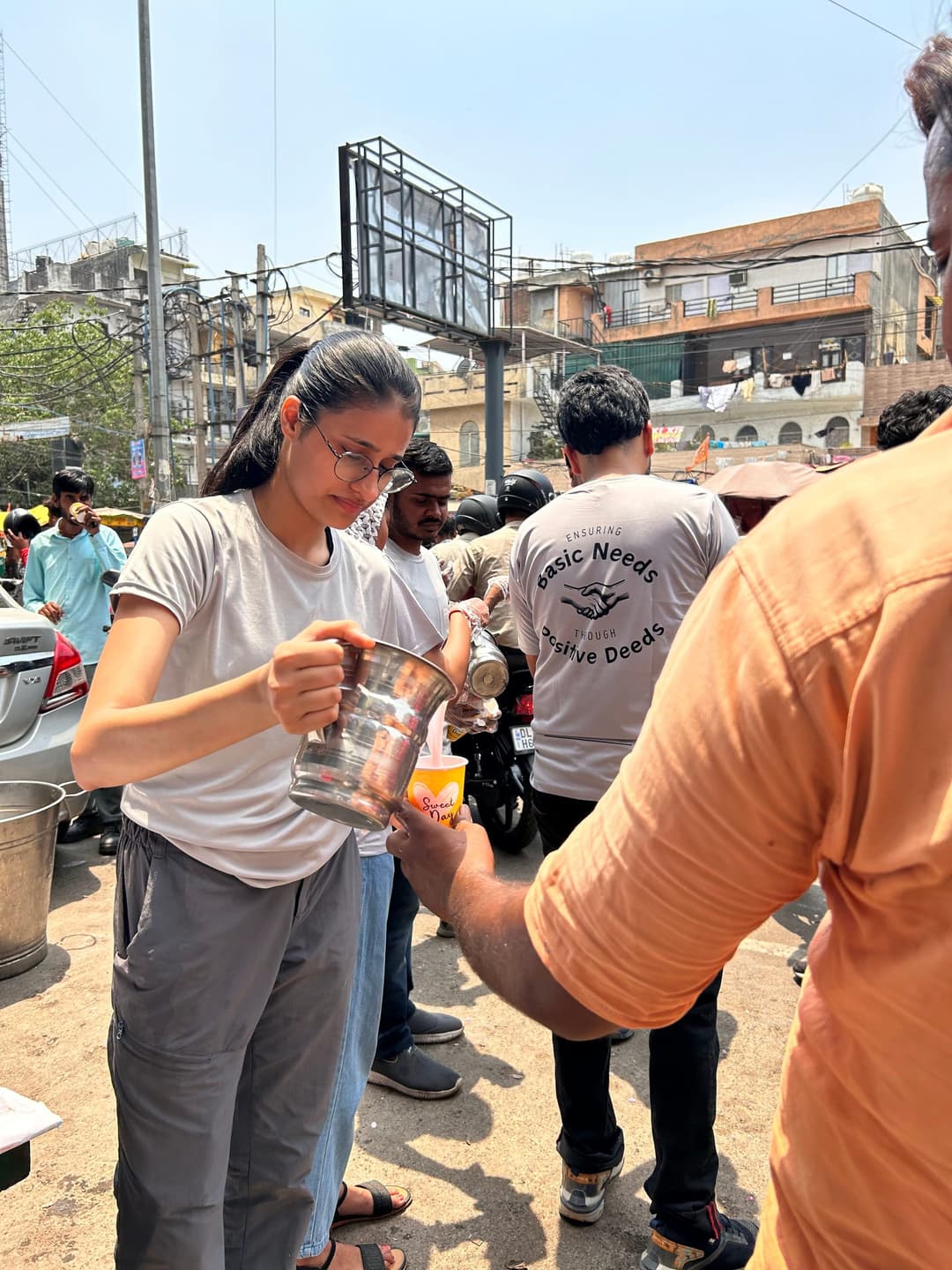 A Helping Hut volunteer in a branded t-shirt pours a cool drink from a steel jug into a cup for a passerby on a busy city street during a summer relief drive.