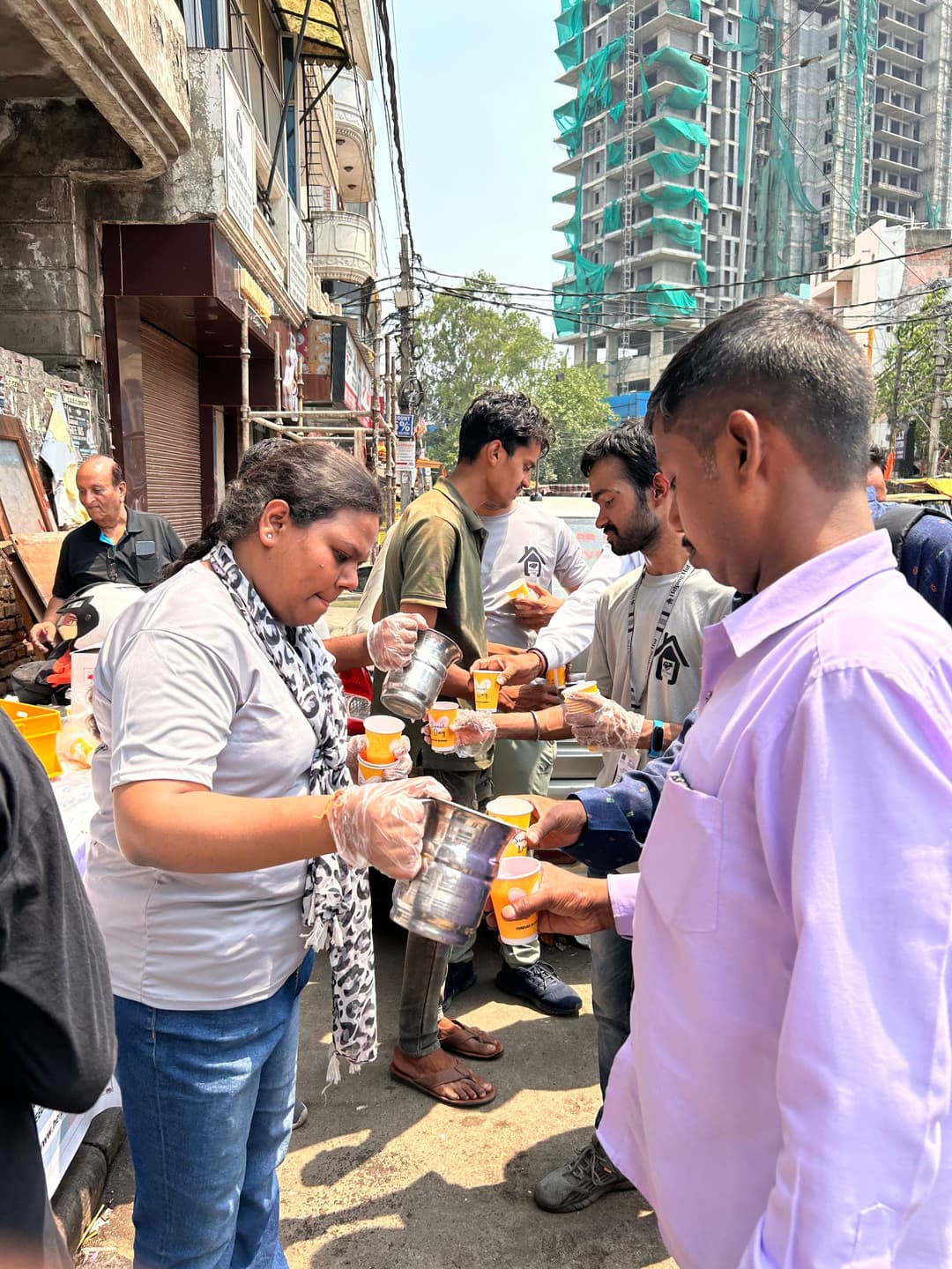 Helping Hut volunteers wearing gloves pour drinks from steel jugs into cups for a line of local residents on a city street.