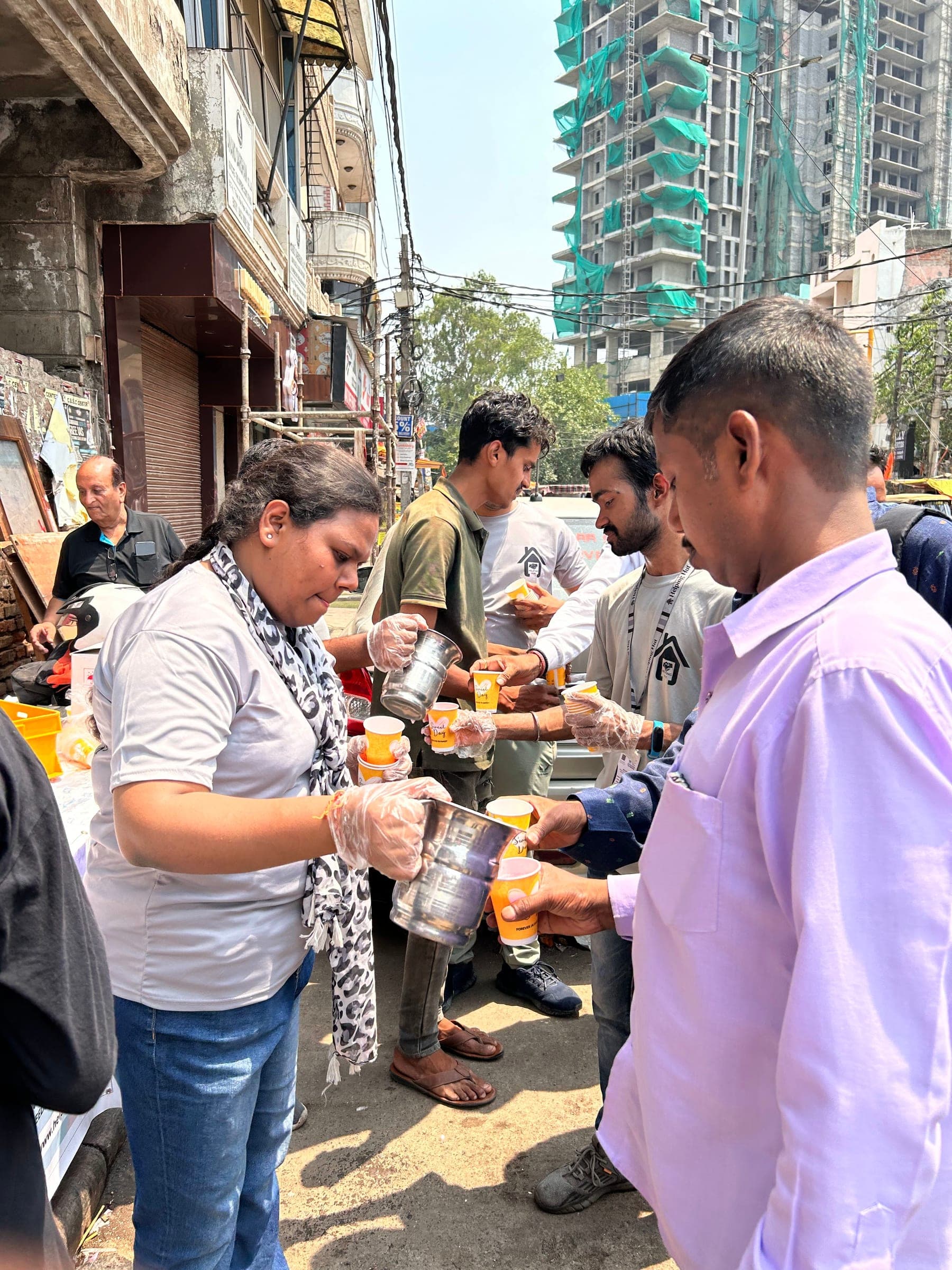 Helping Hut volunteers wearing gloves pour drinks from steel jugs into cups for a line of local residents on a city street.