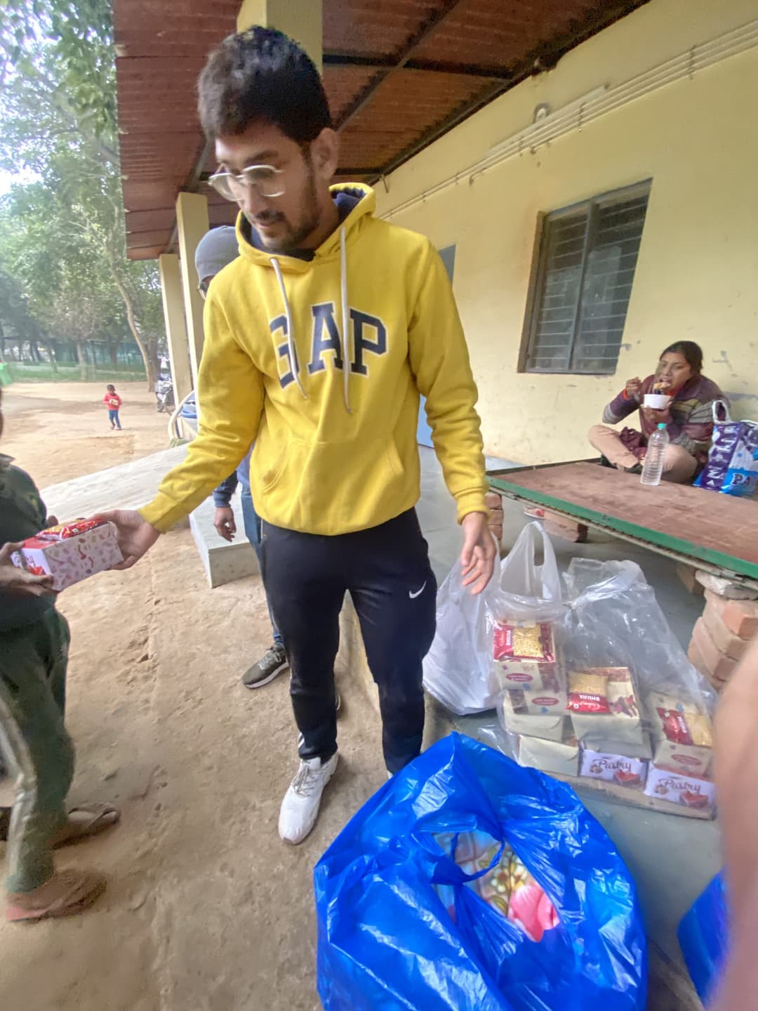 A Helping Hut volunteer in a yellow hoodie hands a packed food box to a child, with bags full of ration boxes waiting on the ground beside a bench.