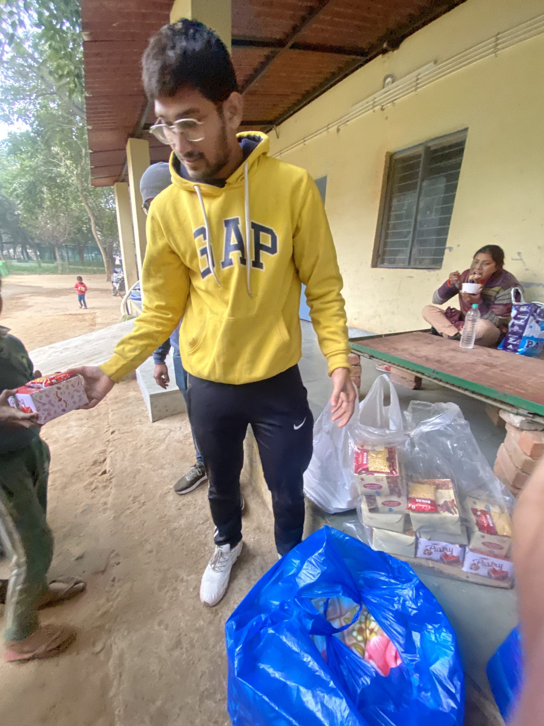 A Helping Hut volunteer in a yellow hoodie hands a packed food box to a child, with bags full of ration boxes waiting on the ground beside a bench.