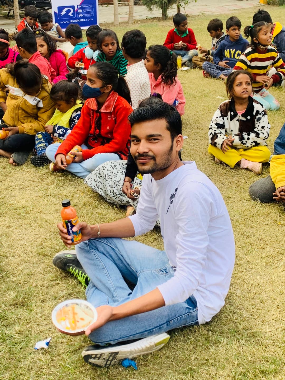 Children sitting on the grass sharing a meal, a volunteer among them