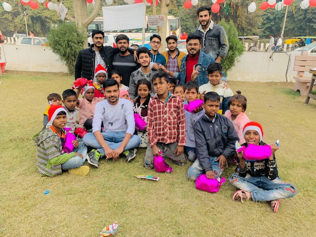 Volunteers sitting on the grass with children holding wrapped gifts, some in Santa hats