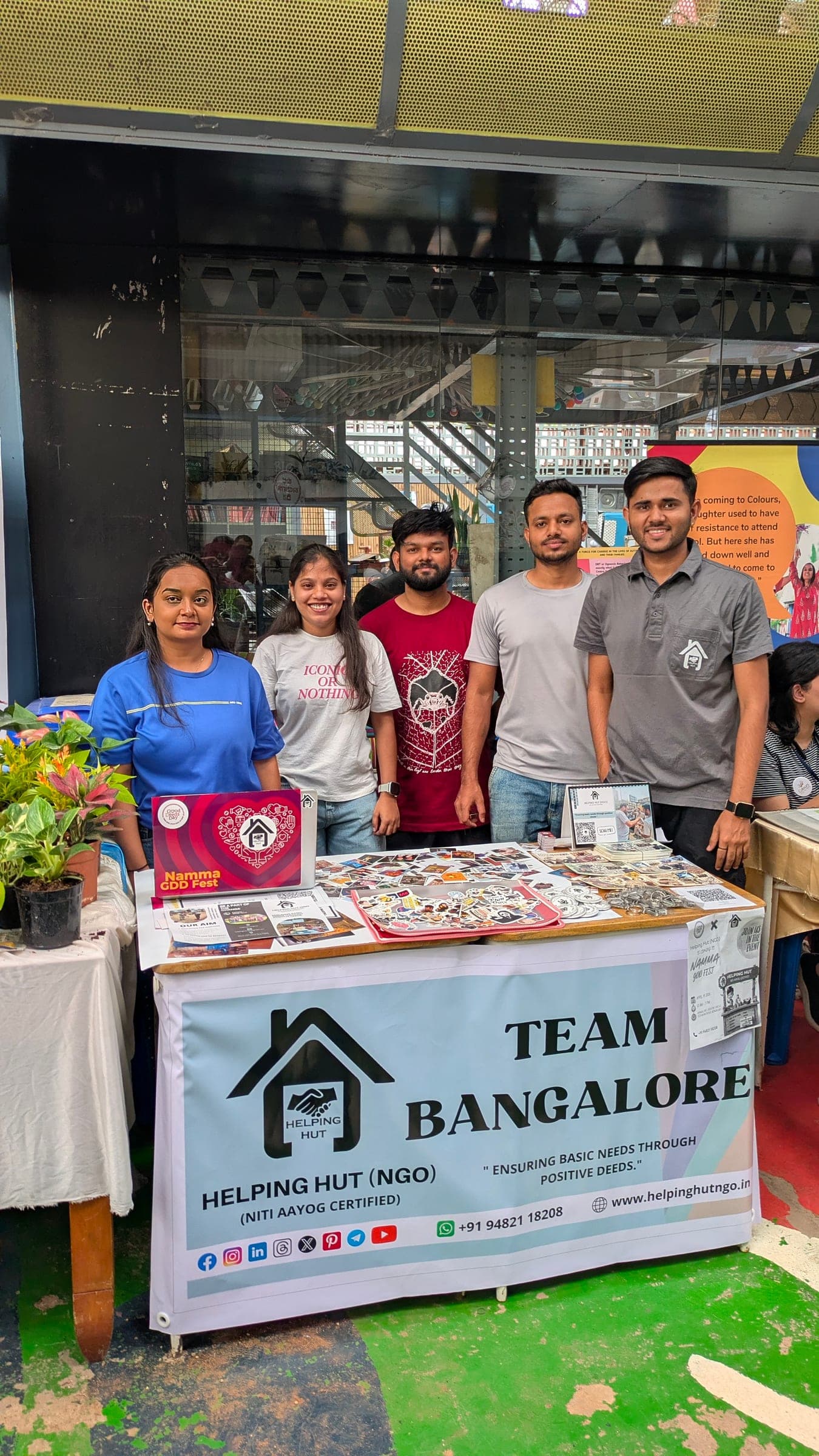 Helping Hut volunteers posing together behind the Team Bangalore stall banner at a community fundraising event