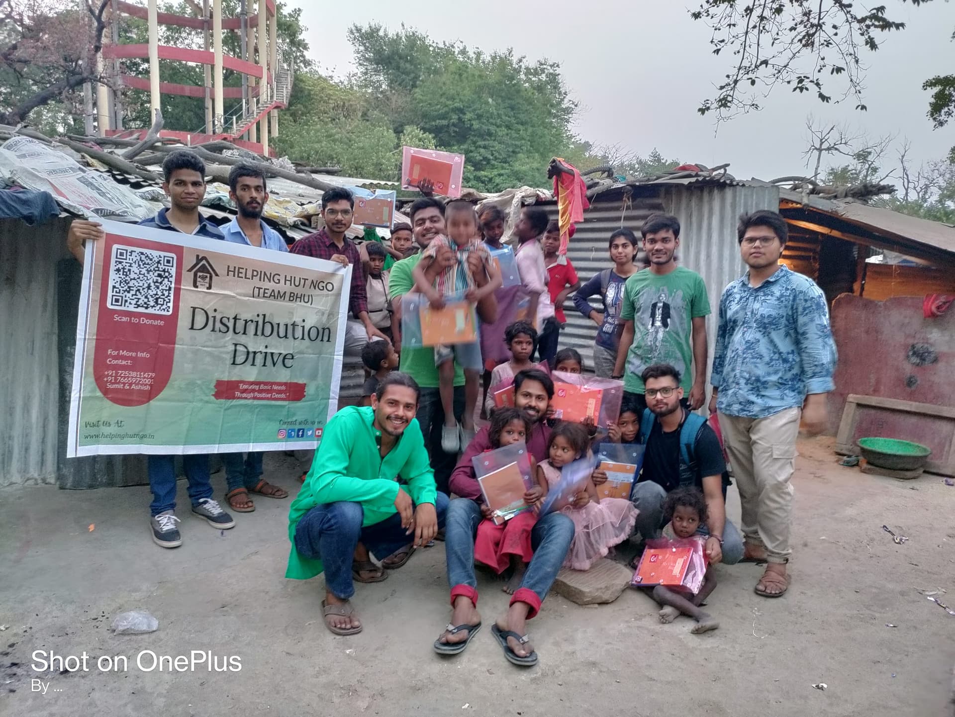 Helping Hut volunteers holding a Team BHU Distribution Drive banner with children receiving supply kits in a Varanasi settlement