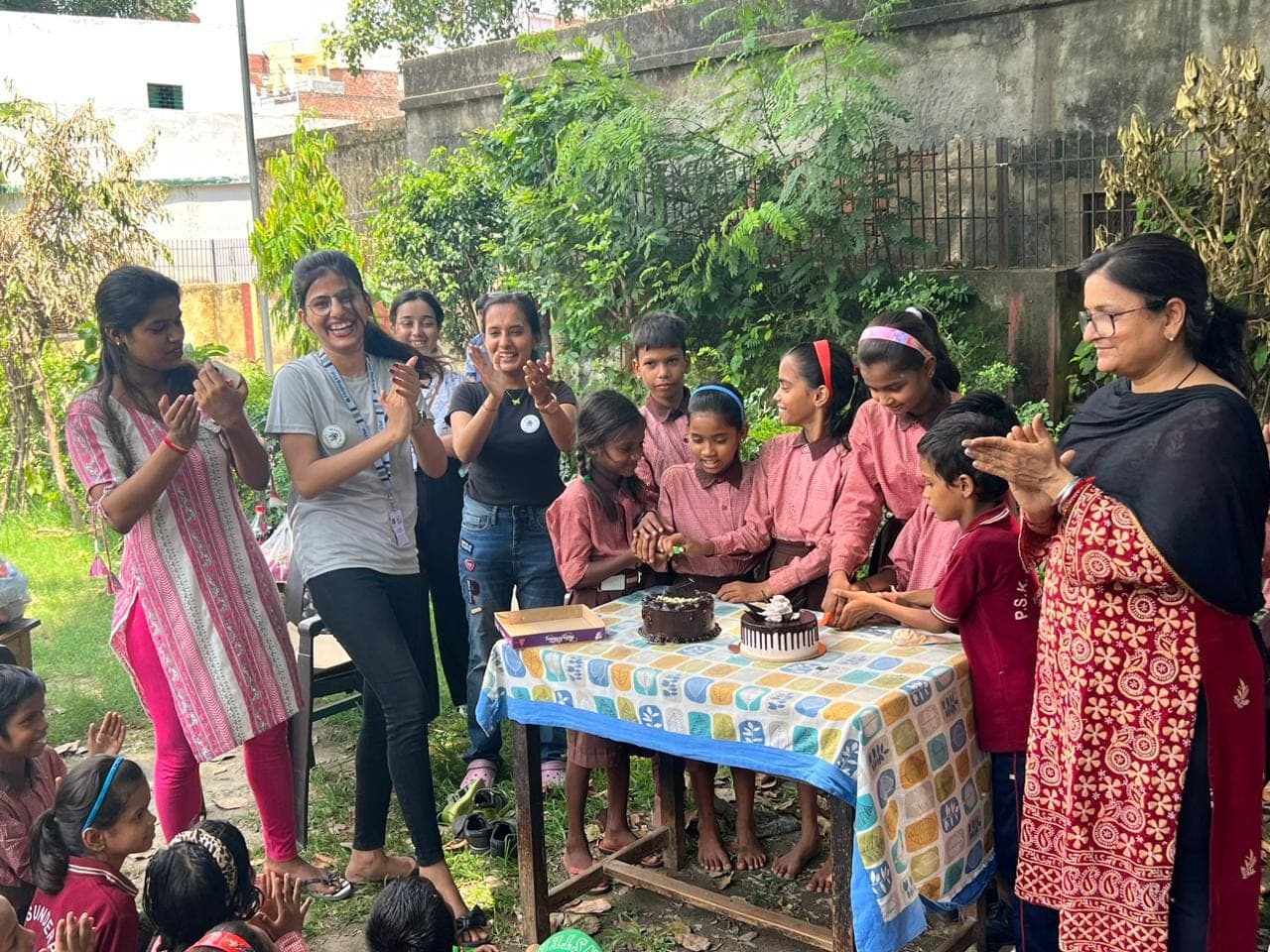Children in pink school uniforms cut chocolate cakes at a decorated table as Helping Hut volunteers clap and cheer around them outdoors.