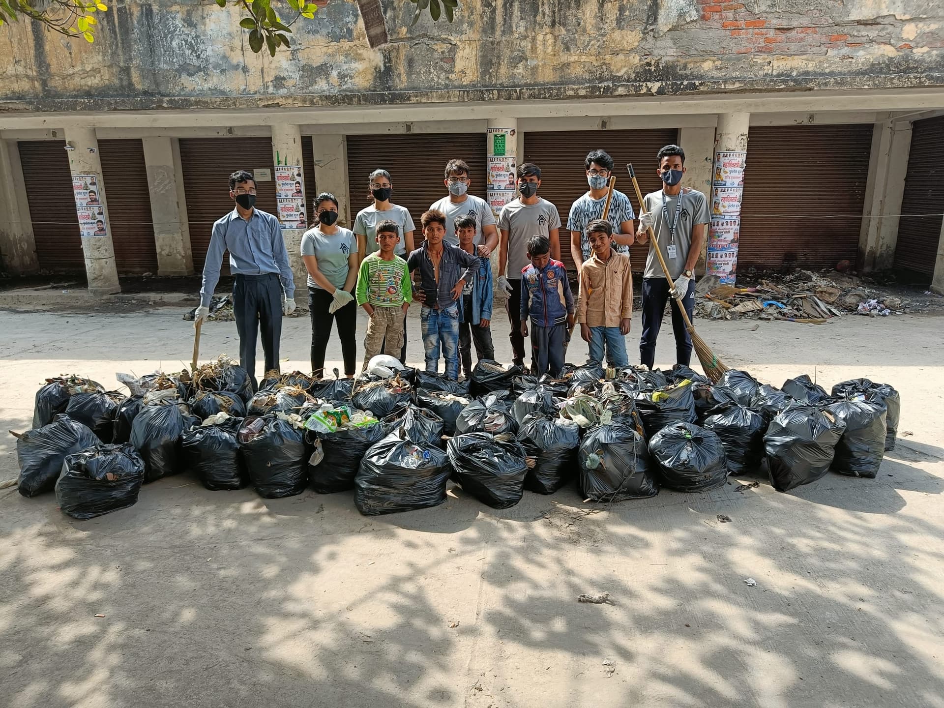 Helping Hut volunteers and local children pose with brooms behind rows of filled black garbage bags on a street they have just cleaned.